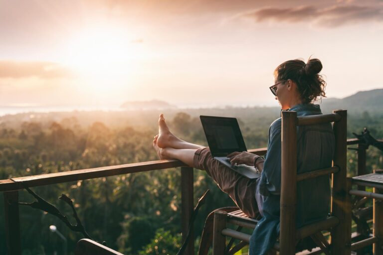 Woman on laptop sitting on deck overlooking a forest and ocean